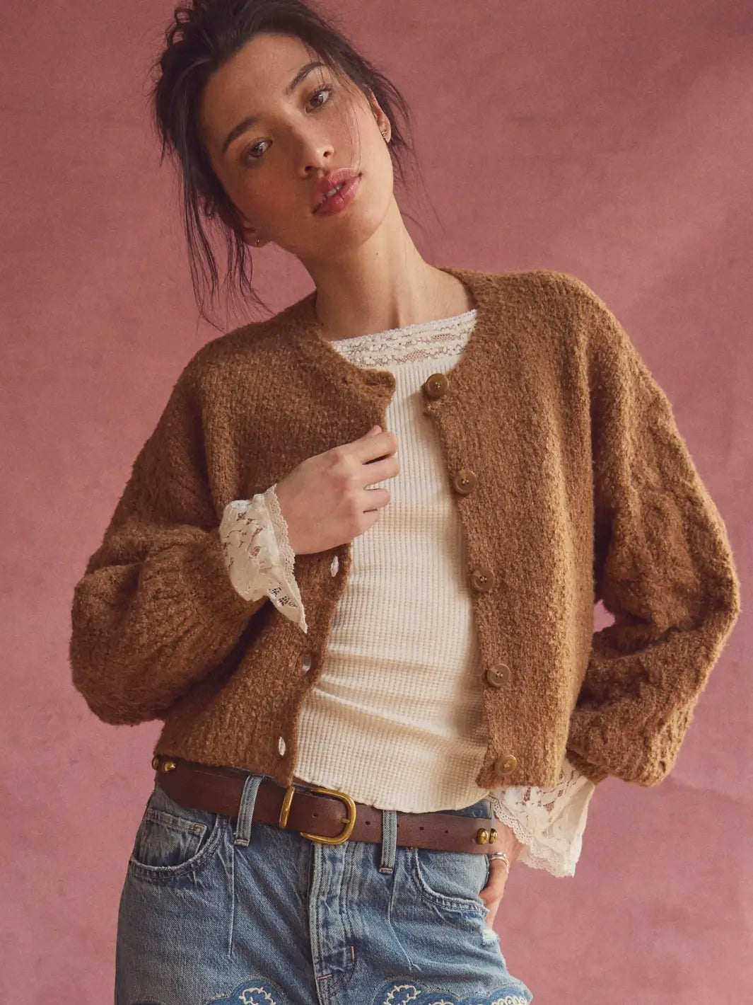 A woman wearing a brown fuzzy button-front Thistle Cardi in Macadamia from Free People over a cream ribbed top, paired with blue jeans and a brown belt, posed against a dusty pink background | Twentyseven Toronto
