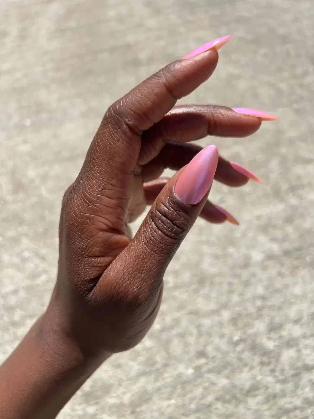 A side profile of a hand with deep skin tone showing off long, pointed nails coated in iridescent pink Death Valley Nails Throwing Spaghetti At The Wall Of Your Heart To See What Sticks Nail Polish | Twentyseven Toronto