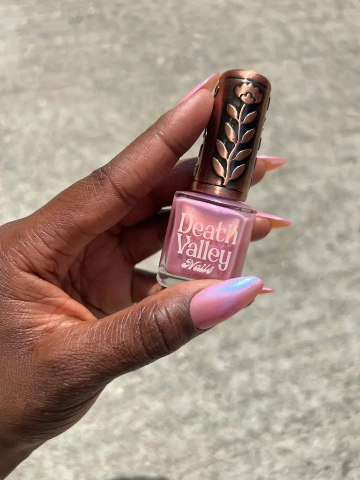 A hand with deep skin tone holds up a bottle of Death Valley Nails Throwing Spaghetti At The Wall Of Your Heart To See What Sticks Nail Polish to display the iridescent pink color on the nails | Twentyseven Toronto