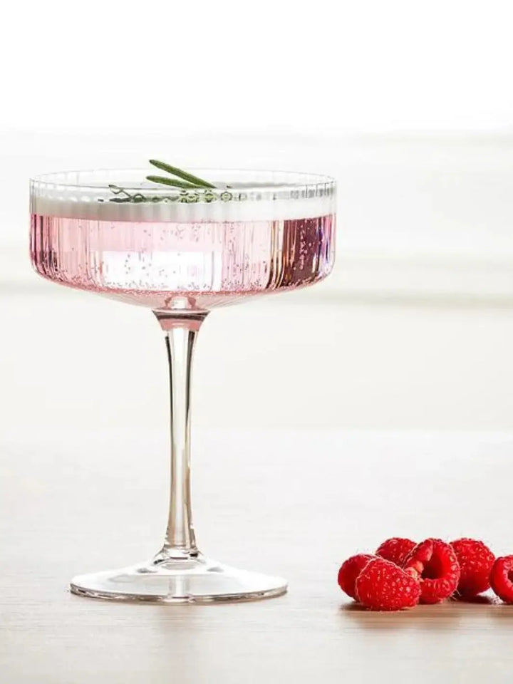 An Abbott Collection Tight Optic Cocktail Coupe filled with a pink drink and garnished with a sprig of rosemary sitting on a table next to fresh raspberries | Twentyseven Toronto