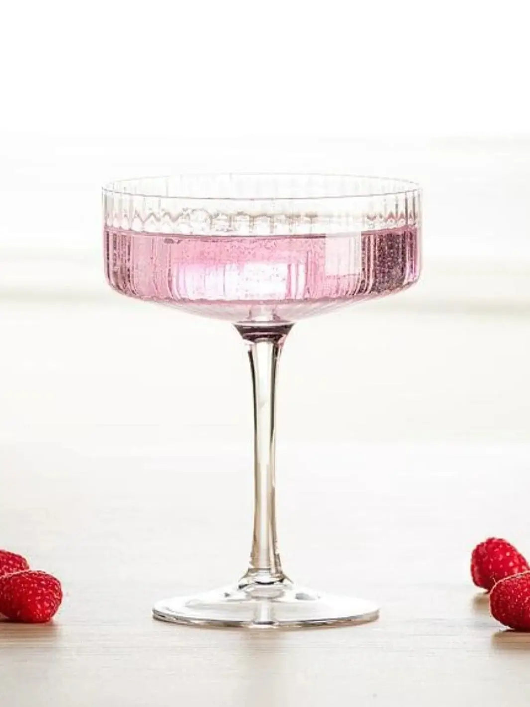 An Abbott Collection Tight Optic Cocktail Coupe holding a bubbly pink beverage, flanked by fresh raspberries on a light-colored surface | Twentyseven Toronto
