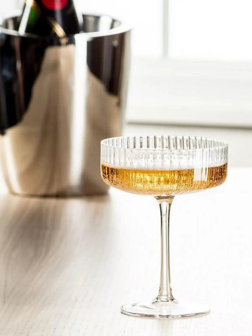 An Abbott Collection Tight Optic Cocktail Coupe filled with a golden, carbonated drink sits on a wooden tabletop with a metallic wine bucket blurred in the background | Twentyseven Toronto