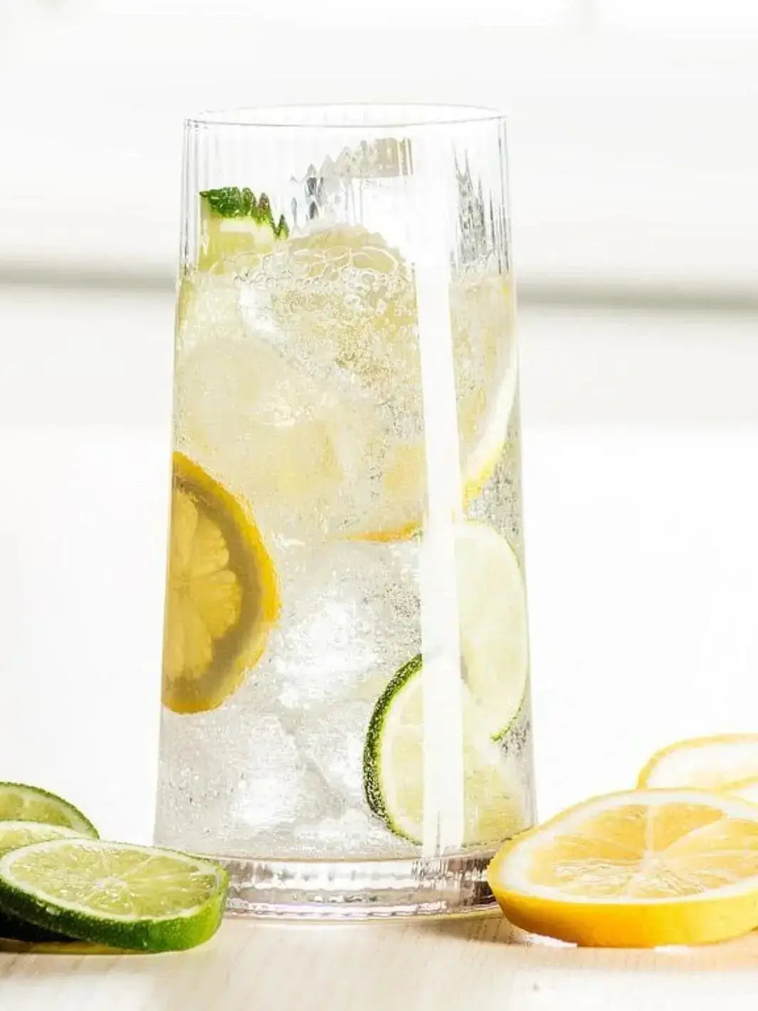 An Abbott Collection Tight Optic Highball glass is filled with a bubbly beverage, ice cubes, and fresh lemon and lime slices on a light tabletop | Twentyseven Toronto