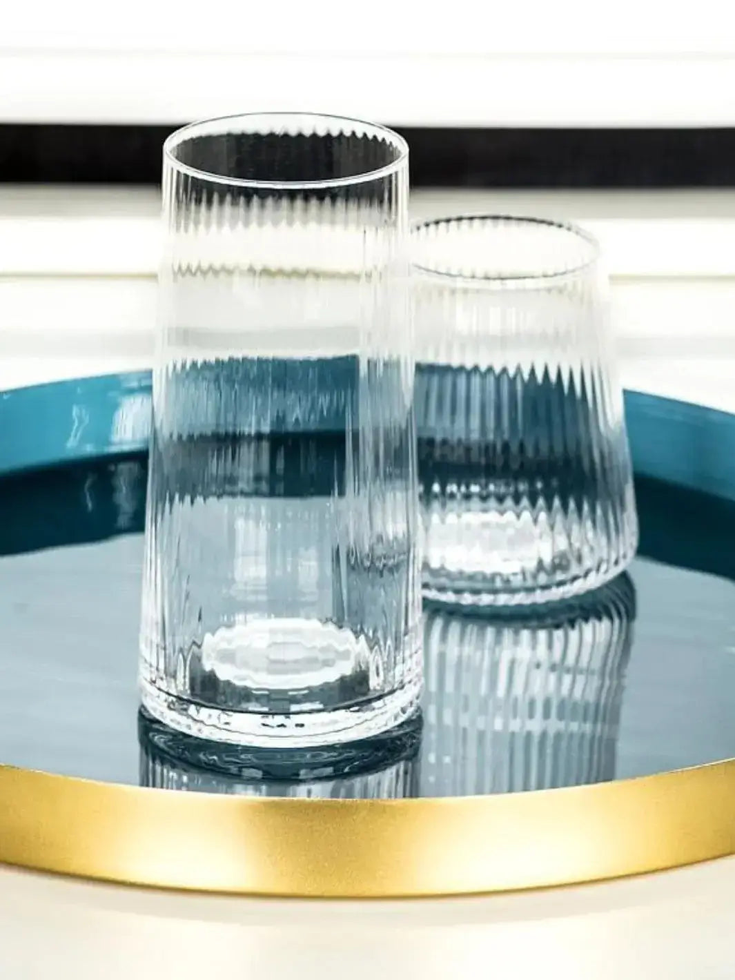An empty, clear glass Abbott Collection Tight Optic Highball sits alongside a matching tumbler on a reflective teal tray with a gold rim | Twentyseven Toronto