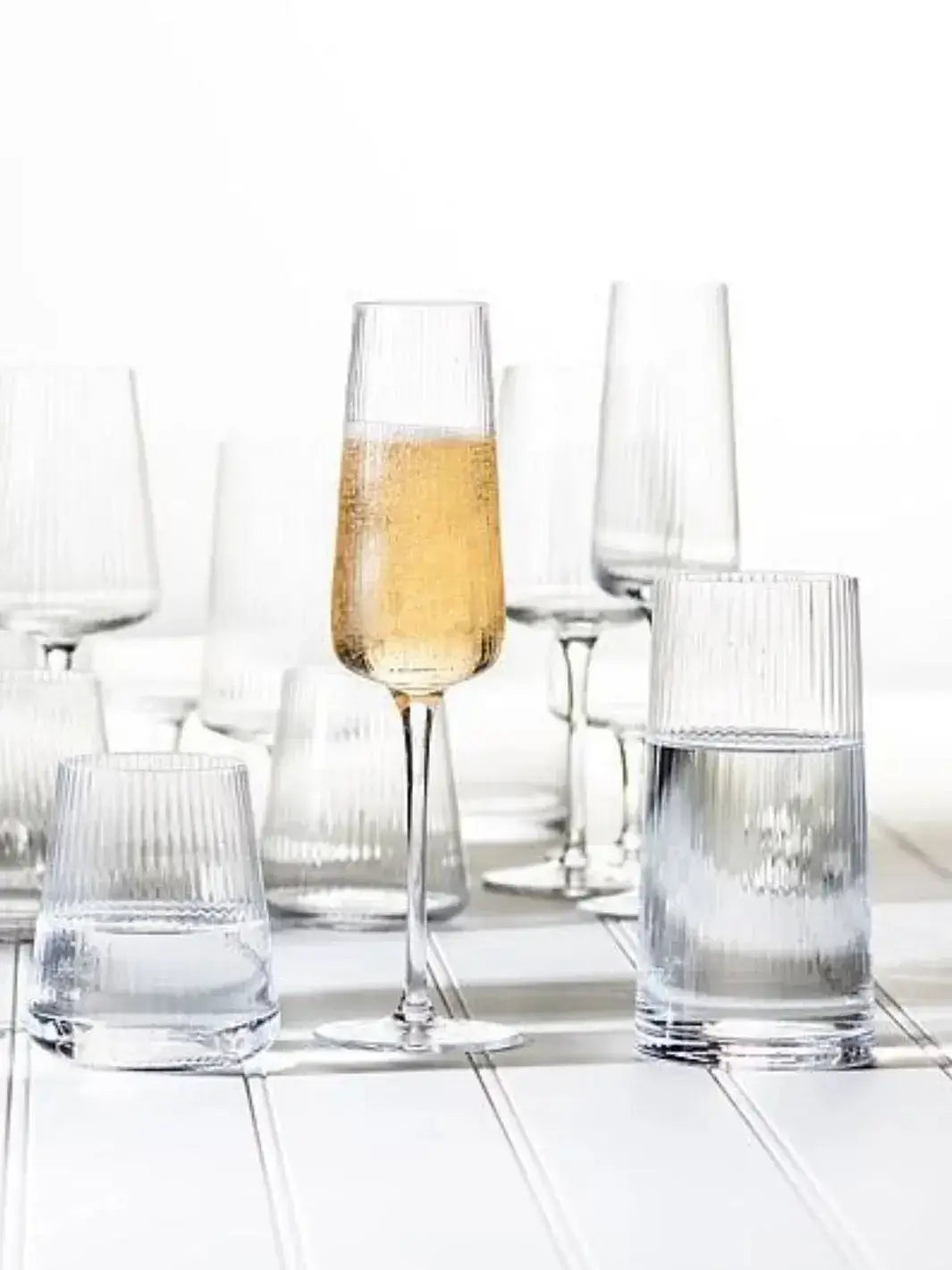An Abbott Collection Tight Optic Highball glass filled with water sits on a white slatted surface amidst a collection of matching ribbed stemware and tumblers | Twentyseven Toronto