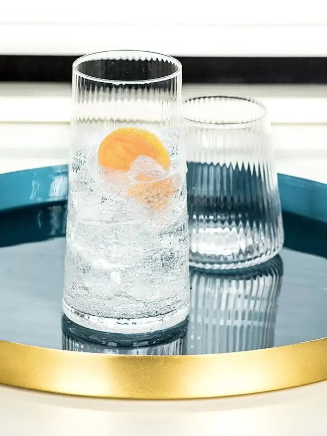 An Abbott Collection Tight Optic Highball glass filled with ice, a citrus slice, and sparkling water sits on a glossy blue and gold tray next to a matching tumbler | Twentyseven Toronto