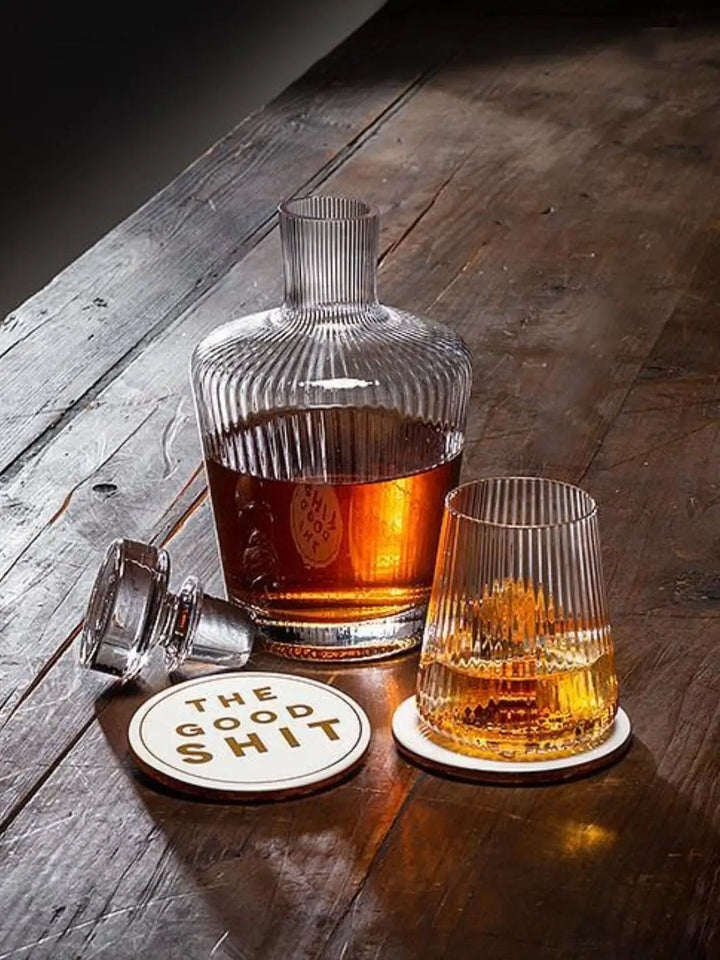 An Abbott Collection Tight Optic Tumbler filled with amber liquor sits on a white coaster on a rustic wooden table next to a matching glass decanter | Twentyseven Toronto