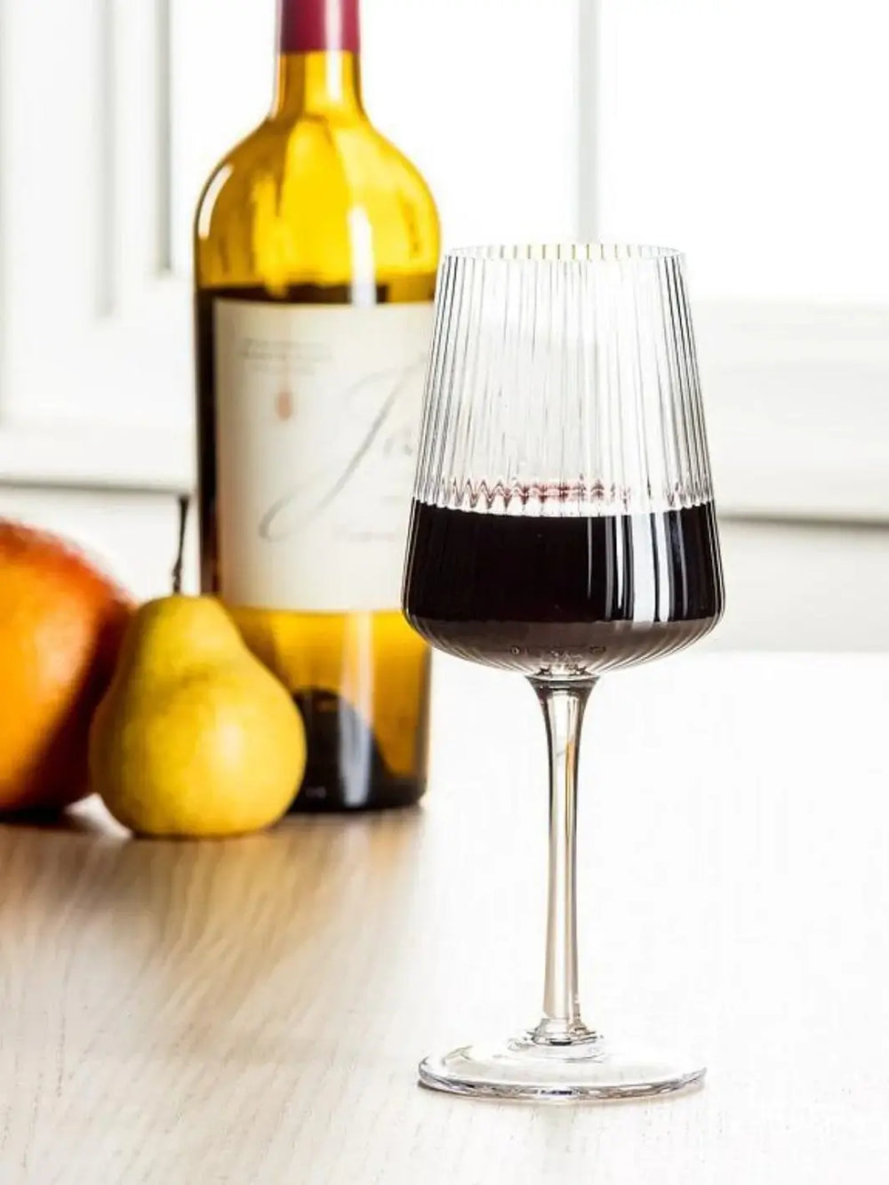An Abbott Collection Tight Optic Wine Glass filled with red wine stands on a light wooden table in front of a whole pear and a wine bottle | Twentyseven Toronto