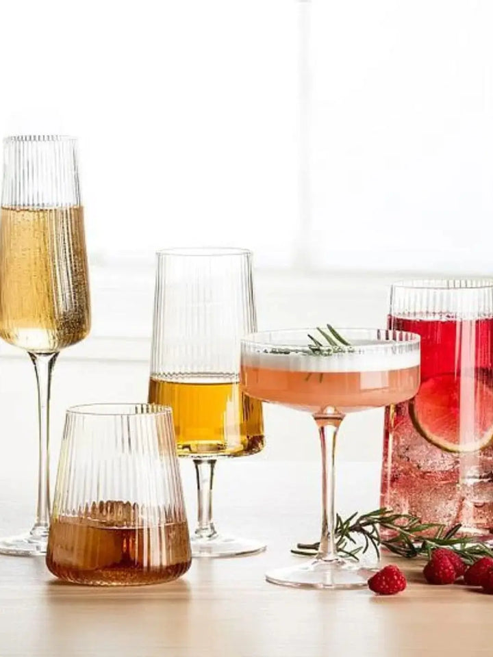 Various ribbed glasses, including an Abbott Collection Tight Optic Wine Glass filled with amber liquid, are arranged on a tabletop alongside a coupe glass and a pitcher with fresh berries | Twentyseven Toronto