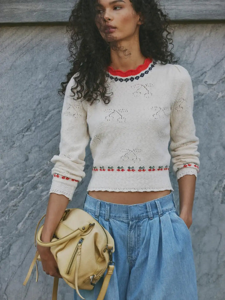 Woman wearing a Free People Tilly Pullover in 'Coconut Milk Combo', a white sweater with red accents and blue jeans, holding a yellow purse against a gray wall | Twentyseven Toronto