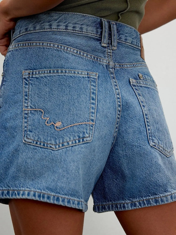 A close-up rear view details the contrast stitching, pocket placement, and embroidered signature wavy line on the back pocket of the medium-wash denim shorts | Twentyseven Toronto