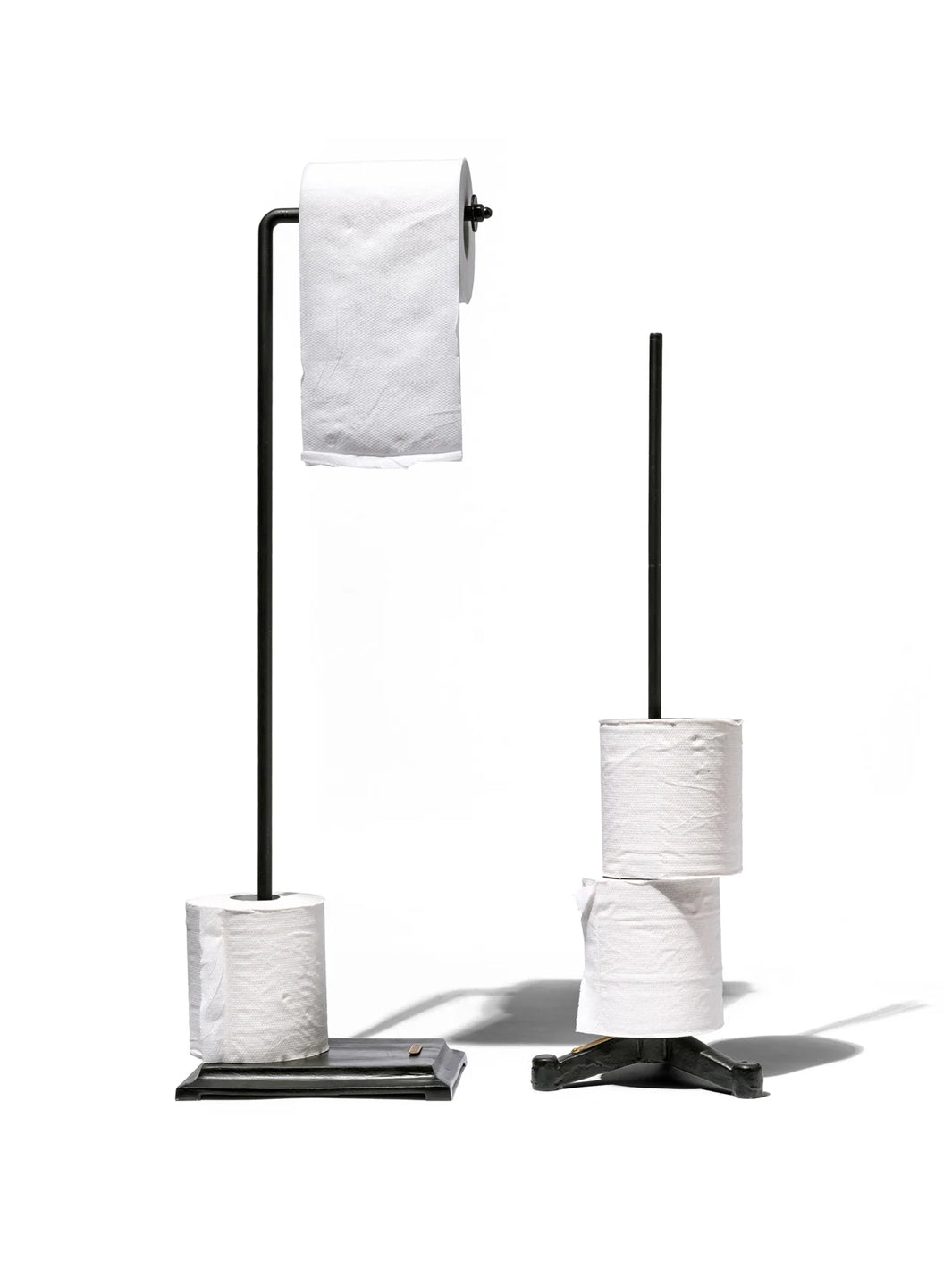 Puebco Angled Toilet Paper Holder in Black, displayed with a toilet paper roll and spare roll on its rectangular iron base, beside a freestanding toilet paper storage stand | Twentyseven Toronto