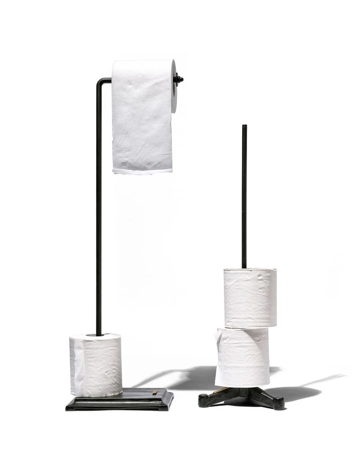 Puebco Angled Toilet Paper Holder in Black, displayed with a toilet paper roll and spare roll on its rectangular iron base, beside a freestanding toilet paper storage stand | Twentyseven Toronto