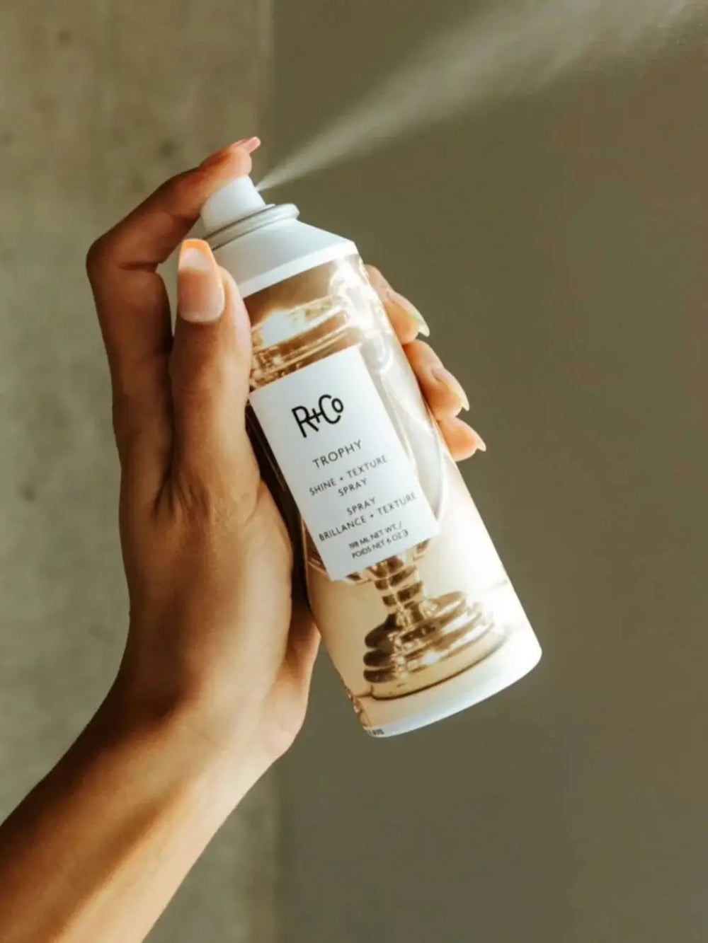A hand with manicured nails dispenses a fine mist from a can of R+Co TROPHY Shine + Texture Spray | Twentyseven Toronto