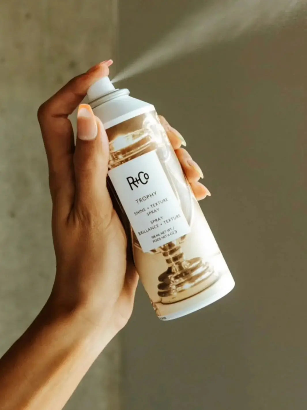 A hand with manicured nails dispenses a fine mist from a can of R+Co TROPHY Shine + Texture Spray | Twentyseven Toronto