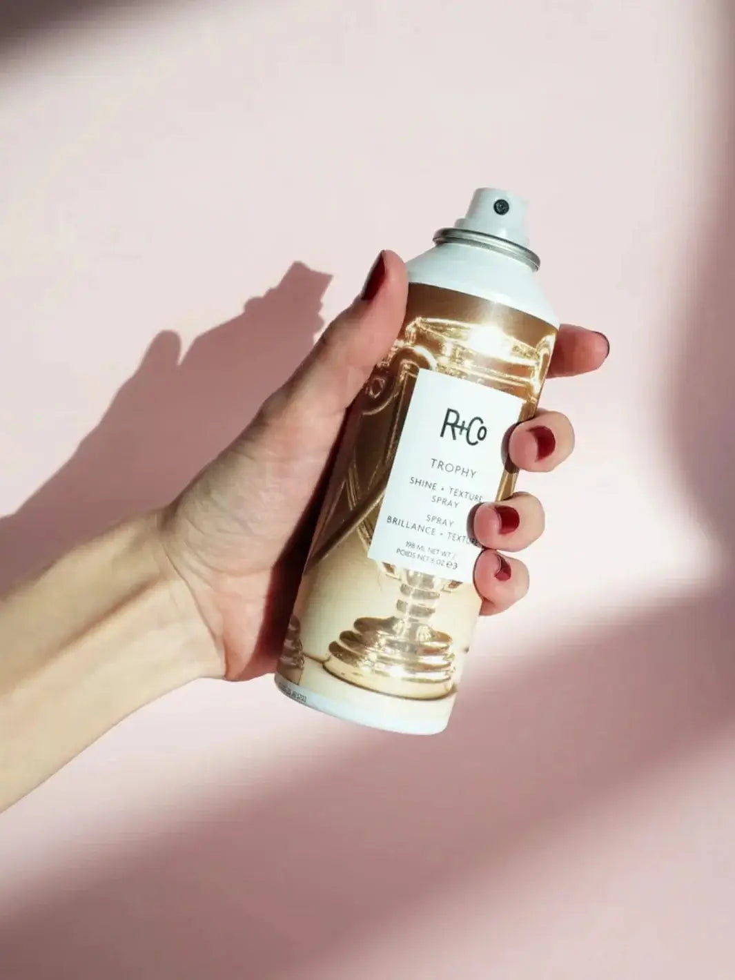 A hand with red fingernails holds a can of R+Co TROPHY Shine + Texture Spray against a soft pink background under casting shadows | Twentyseven Toronto