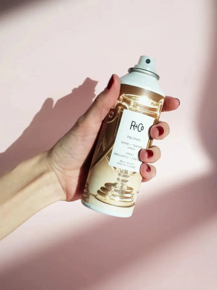 A hand with red fingernails holds a can of R+Co TROPHY Shine + Texture Spray against a soft pink background under casting shadows | Twentyseven Toronto