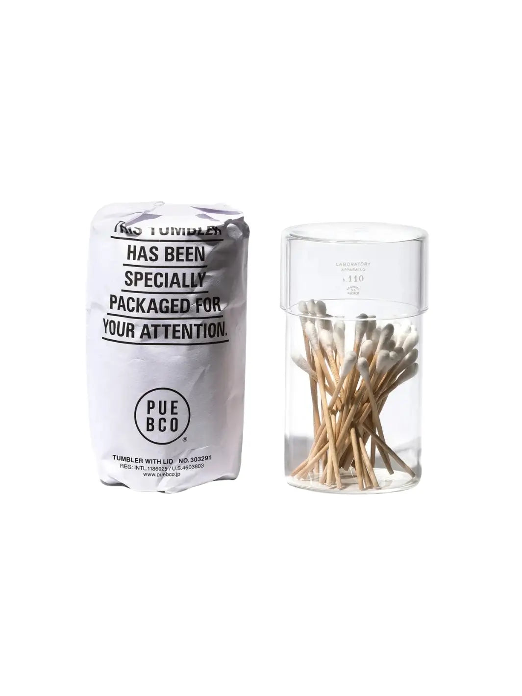 A side-by-side view showing the clear glass Tumbler With Lid from Puebco filled with cotton swabs next to its branded, white paper protective wrapping | Twentyseven Toronto