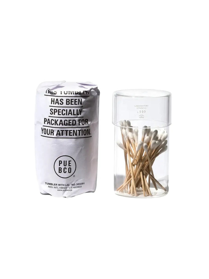 A side-by-side view showing the clear glass Tumbler With Lid from Puebco filled with cotton swabs next to its branded, white paper protective wrapping | Twentyseven Toronto