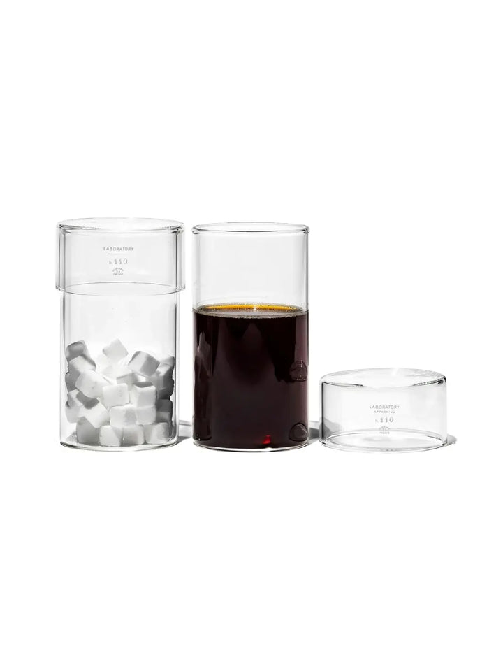 A lifestyle demonstration of the versatile Tumbler With Lid from Puebco, showing one unit filled with sugar cubes, another filled with a dark liquid, and its removable glass lid resting alongside | Twentyseven Toronto