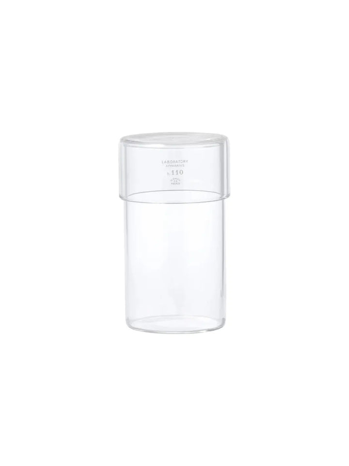 A front-facing product shot of the empty clear glass Tumbler With Lid from Puebco against a stark white backdrop, displaying its minimalist laboratory-inspired branding | Twentyseven Toronto