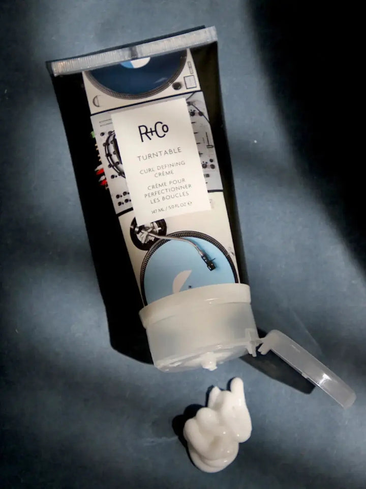 A squeeze tube of R+Co TURNTABLE Curl Defining Cream lies flat with an open cap next to a small dollop of the white styling cream on a dark background | Twentyseven Toronto