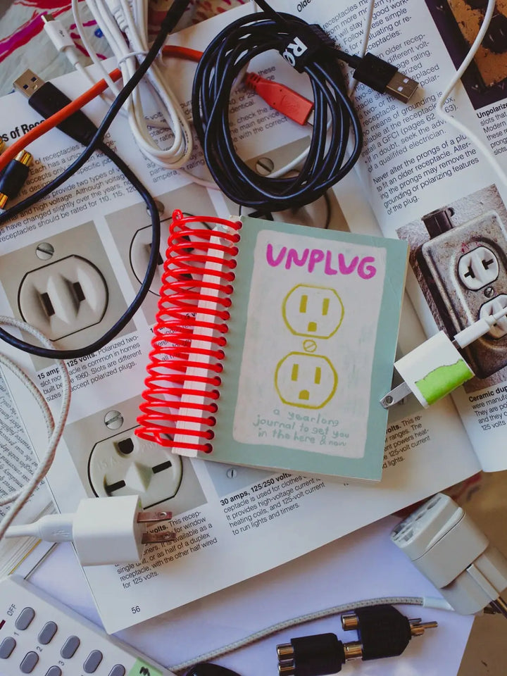 The Unplug Journal from People I've Loved sits on an open book about electrical wiring, surrounded by tangled USB cords and power plugs | Twentyseven Toronto