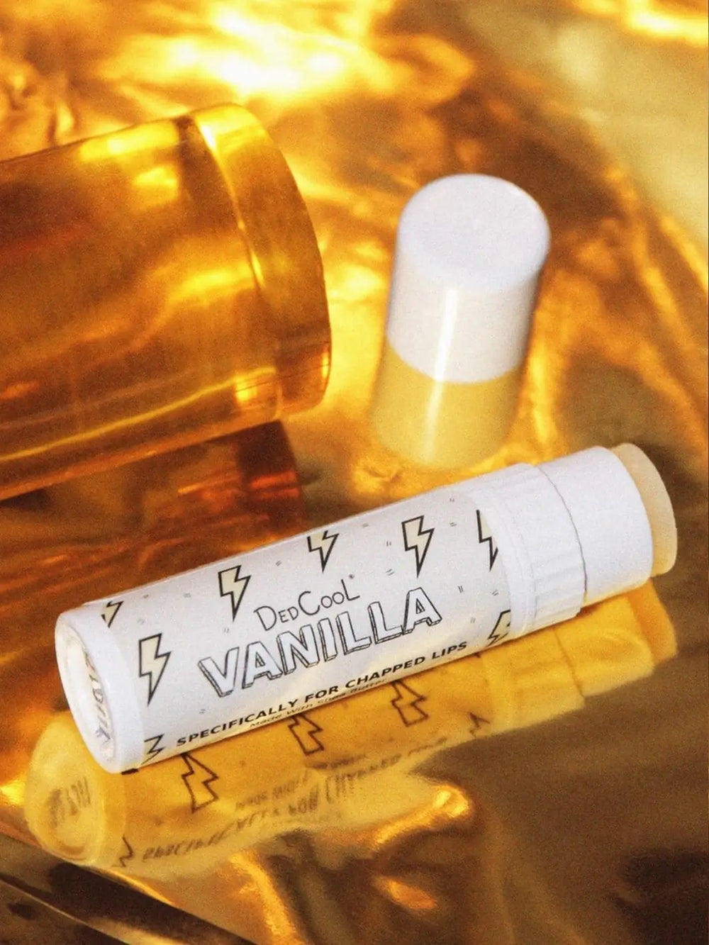 A white tube of Dedcool Vanilla Balm Stick is displayed horizontally on a reflective gold surface next to an amber pill bottle and a white cap | Twentyseven Toronto