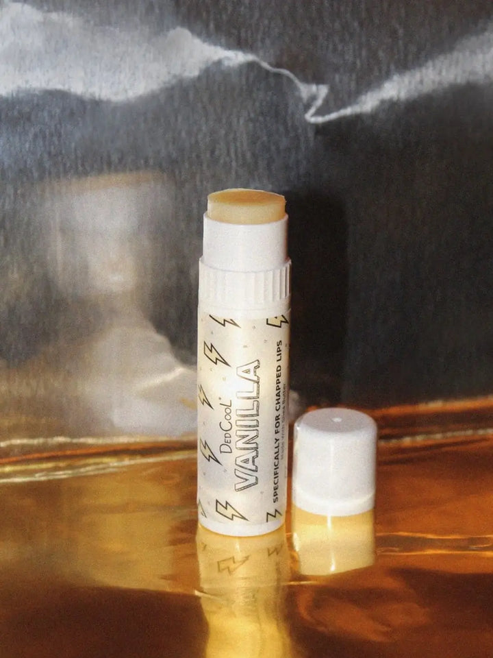 An uncapped tube of Dedcool Vanilla Balm Stick stands on a metallic gold surface against a dark, atmospheric background | Twentyseven Toronto