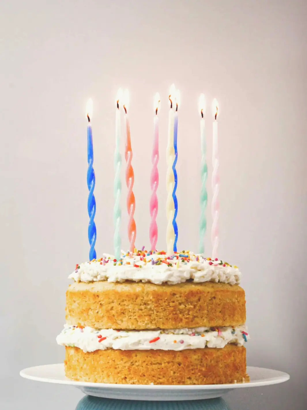 A two-layered cake made from the Glazed Cake Co Vanilla Bean signature kit features fluffy white frosting, colorful sprinkles, and seven lit, spiral birthday candles | Twentyseven Toronto
