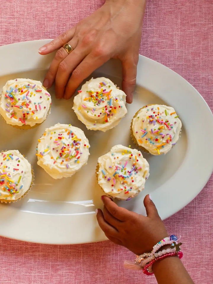 A top-down view shows an adult and a child reaching for frosted vanilla cupcakes decorated with rainbow sprinkles, made using the Glazed Cake Co Vanilla Bean mix | Twentyseven Toronto
