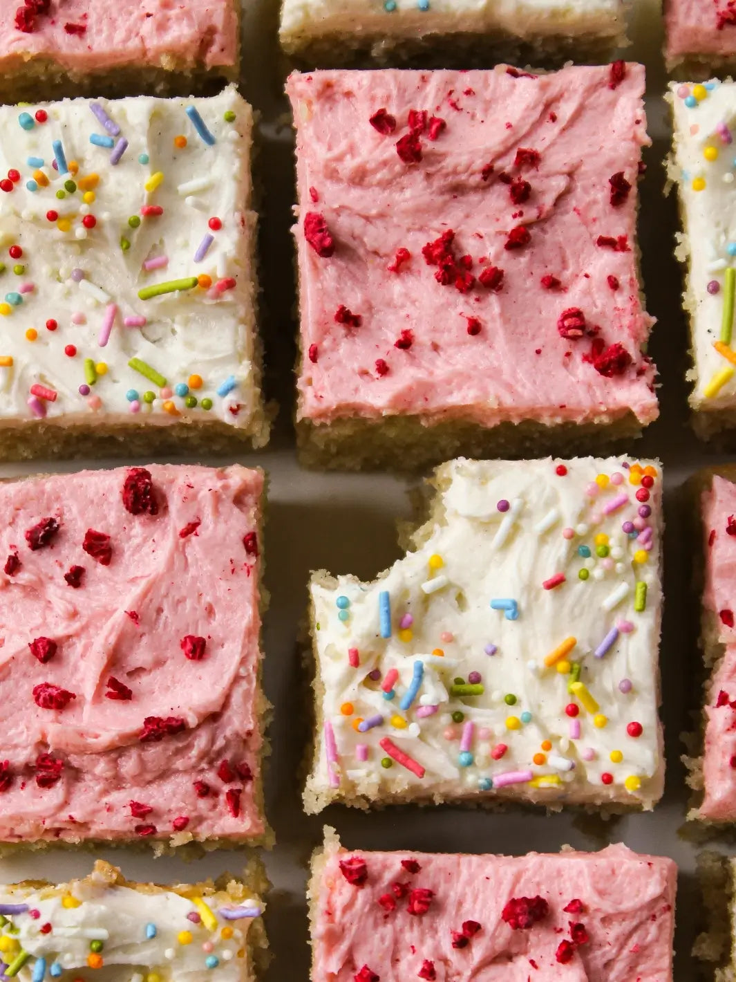 A close-up shot reveals a grid of square cake slices frosted with pink raspberry and white vanilla icing, including a white piece with rainbow sprinkles from the Glazed Cake Co Vanilla Bean kit that has a bite taken out of it | Twentyseven Toronto