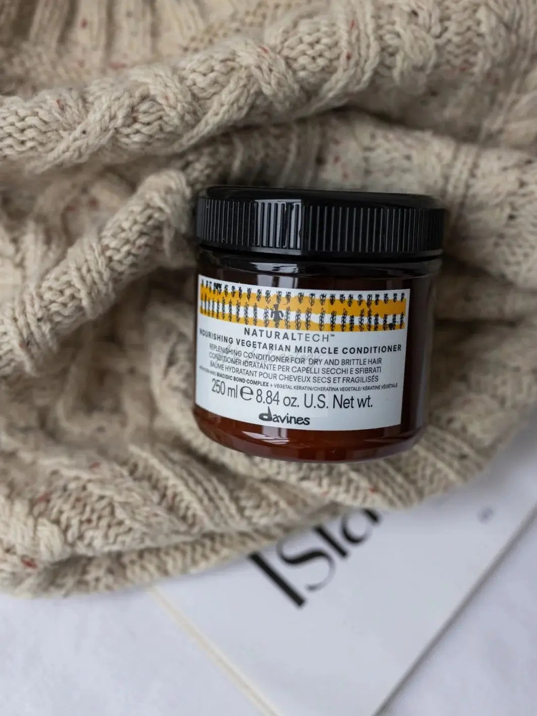 A jar of Davines NOURISHING Vegetarian Miracle Conditioner on top of a textured, beige knit sweater next to a magazine | Twentyseven Toronto