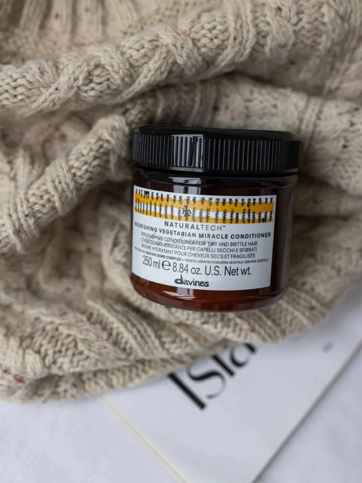 A jar of Davines NOURISHING Vegetarian Miracle Conditioner on top of a textured, beige knit sweater next to a magazine | Twentyseven Toronto