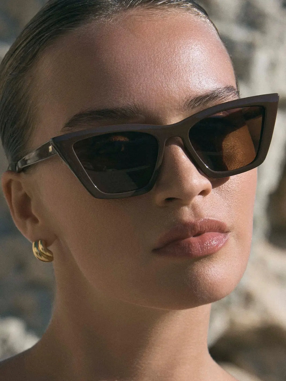 A close up shot of a woman looking wearing thick, angular Le Specs Velodrome - Cocoa sunglasses with dark lenses in an outdoor setting | Twentyseven Toronto