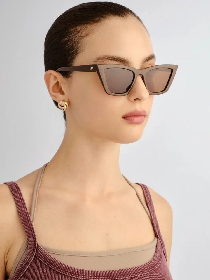 A portrait of a woman looking off to the side, modeling the Le Specs Velodrome - Cocoa sunglasses paired with a twisted gold stud earring | Twentyseven Toronto