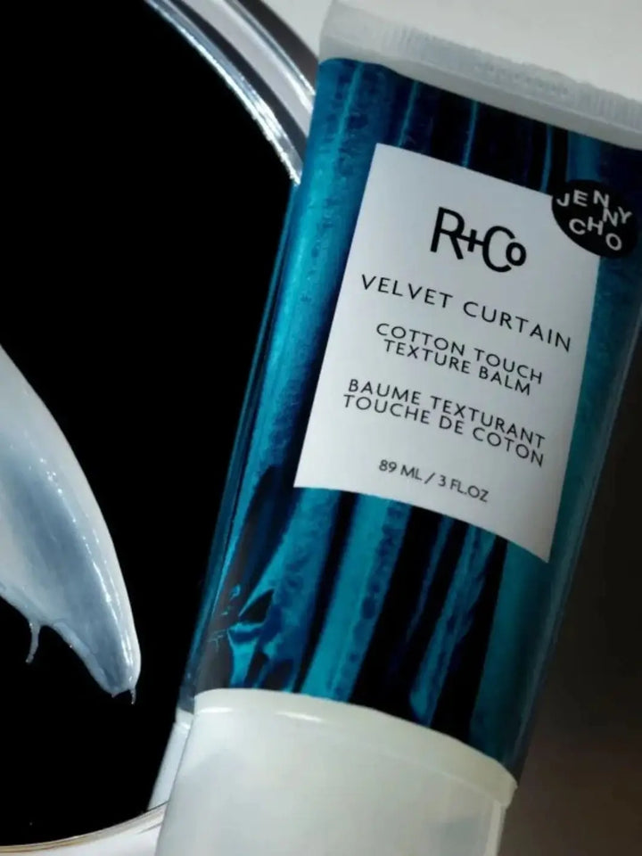 A tube of R+Co VELVET CURTAIN Texture Balm is displayed diagonally in a close-up shot next to a dollop of creamy product against a dark background | Twentyseven Toronto