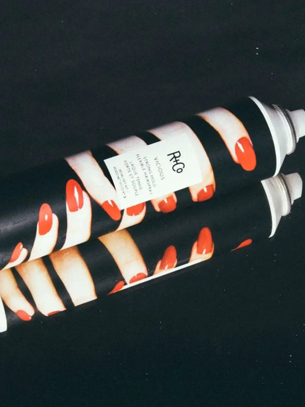 Two aerosol cans of R+Co VICIOUS Strong Hold Flexible Hairspray, featuring a graphic design of fingers with red nails, are stacked horizontally against a solid black background | Twentyseven Toronto