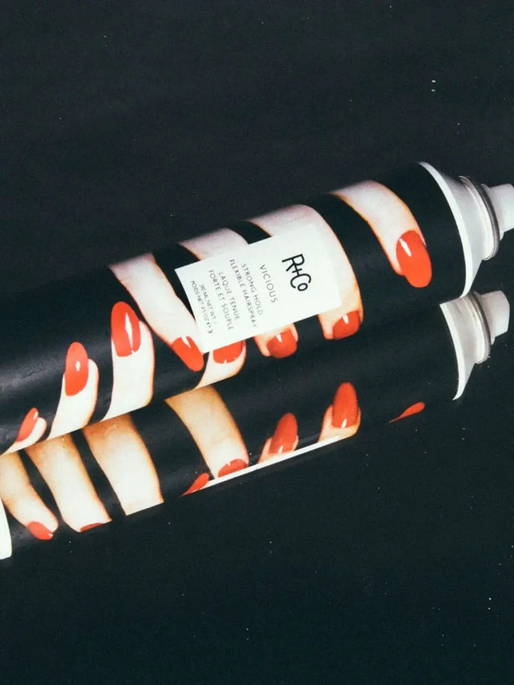 Two aerosol cans of R+Co VICIOUS Strong Hold Flexible Hairspray, featuring a graphic design of fingers with red nails, are stacked horizontally against a solid black background | Twentyseven Toronto