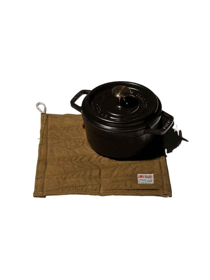 A heavy black cast-iron pot rests on top of a tan canvas Vintage Bivouac Trivet from Puebco against a transparent background | Twentyseven Toronto