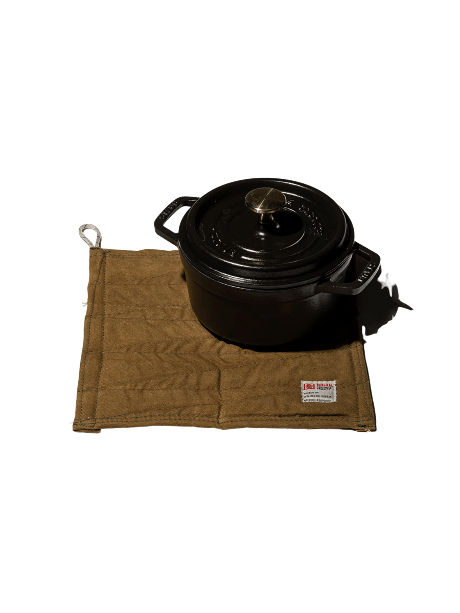A heavy black cast-iron pot rests on top of a tan canvas Vintage Bivouac Trivet from Puebco against a transparent background | Twentyseven Toronto