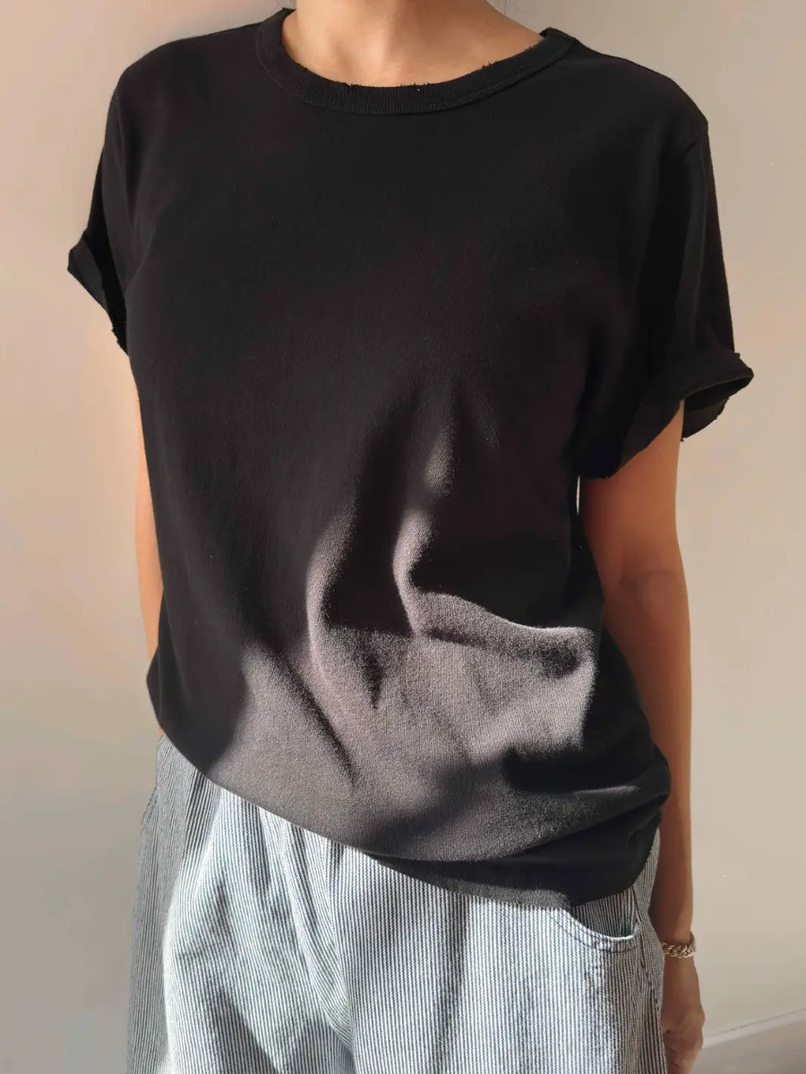 A person models the black relaxed-fit Le Bon Shoppe Vintage Boy Tee - True Black from a slight angle, highlighting the rolled short sleeves and a subtly distressed neckline | Twentyseven Toronto
