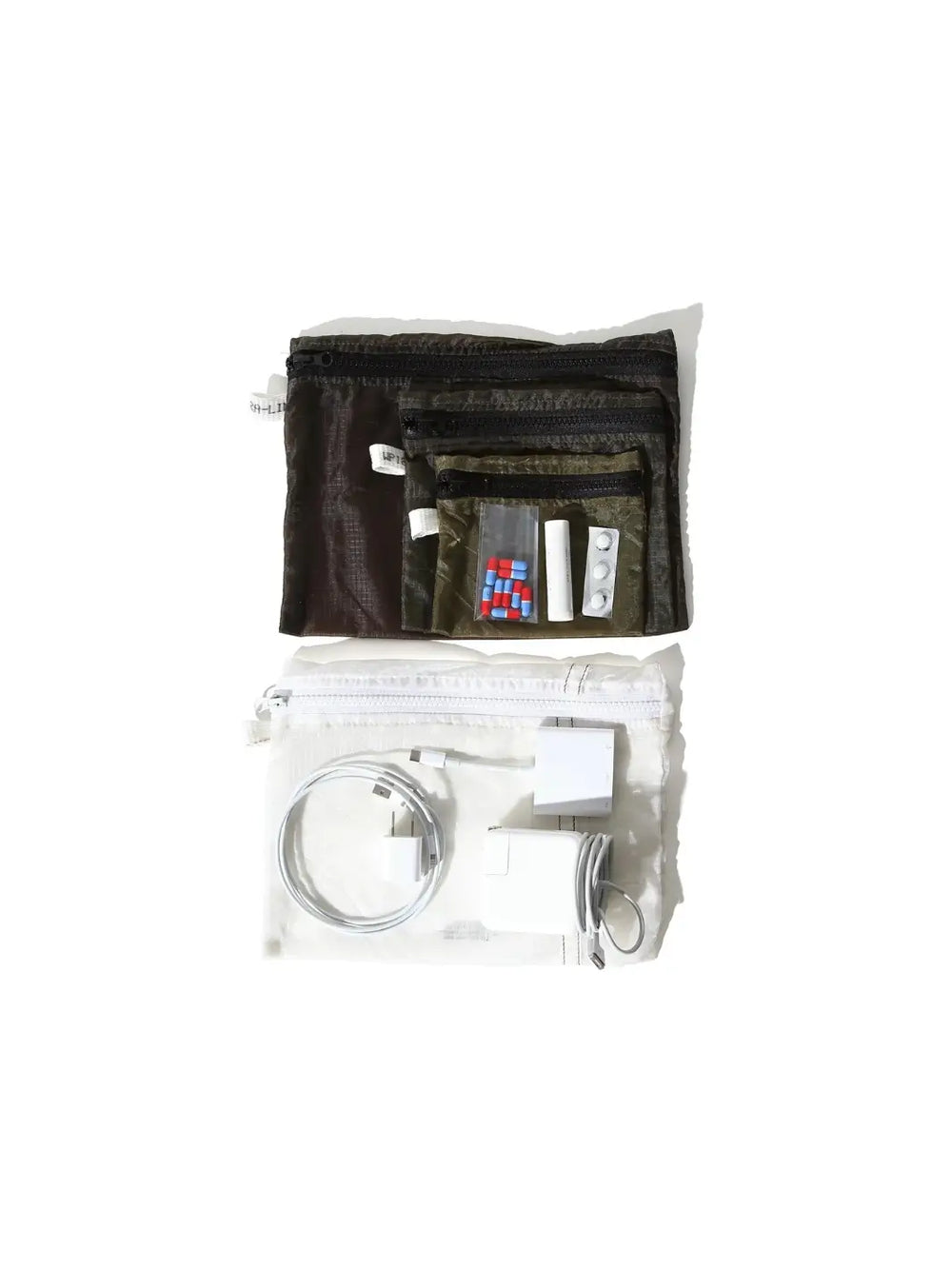 Everyday travel essentials, including charging blocks and medication organized inside and around the semi-translucent, white Vintage Parachute Light Pouch Medium from Puebco alongside a nested set of smaller olive-green pouches | Twentyseven Toronto