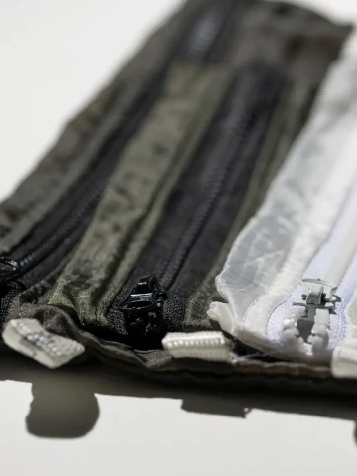 A close-up, angled shot highlights the durable zippered closures and crinkled nylon material of a white Vintage Parachute Light Pouch Medium from Puebco resting next to layered, olive green pouches | Twentyseven Toronto