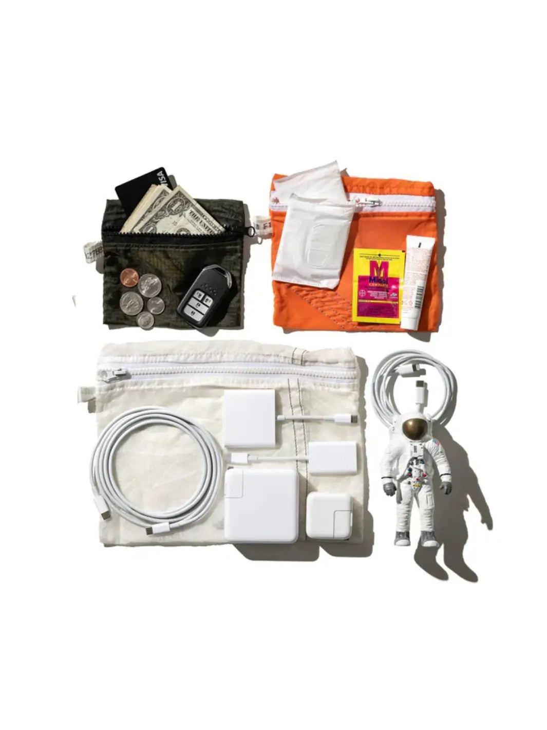 A top-down flat lay displays everyday items organized in Vintage Parachute Light Pouches from Puebco alongside an astronaut figurine, personal care items on an orange pouch, and cash and keys on a dark green pouch | Twentyseven Toronto