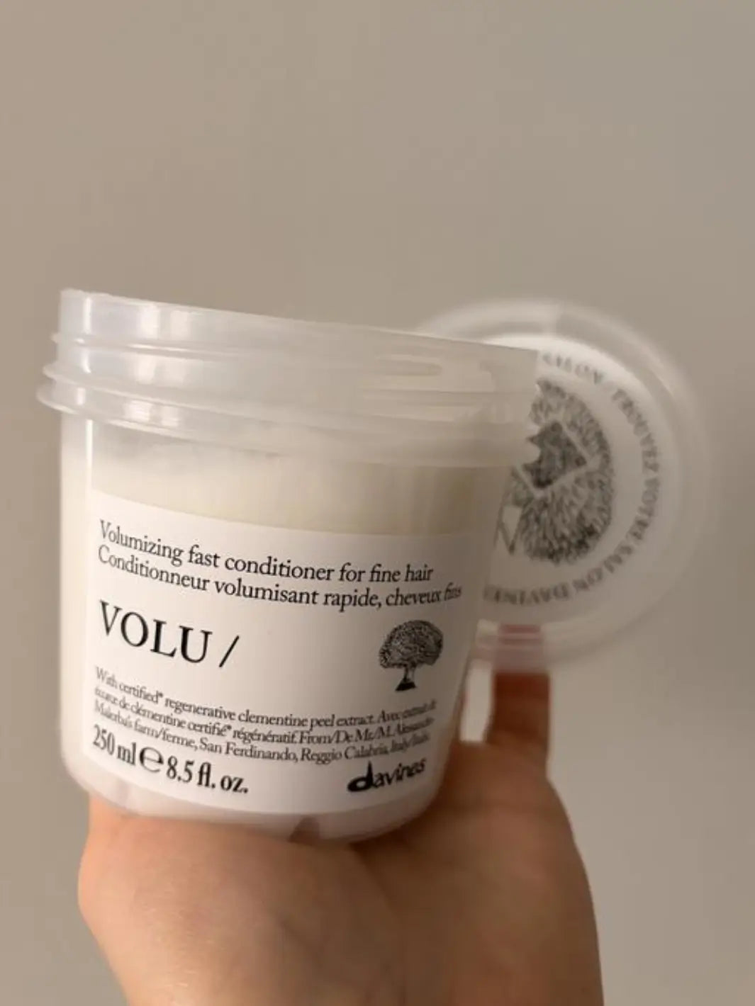 A close-up shot shows a hand holding an open 250 ml tub of Davines VOLU Conditioner for fine hair with the translucent lid resting in the background | Twentyseven Toronto