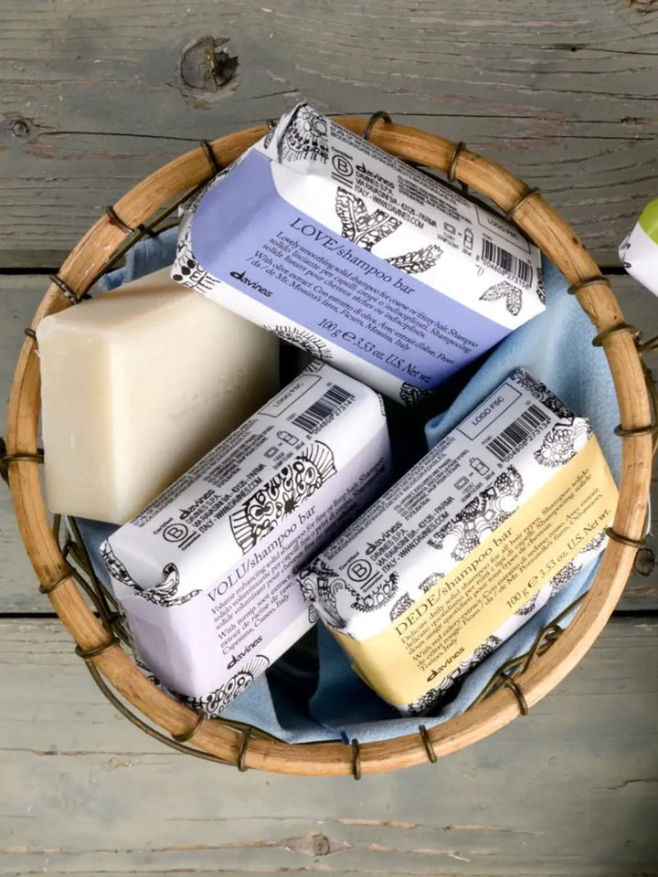 A round woven basket holds an unwrapped bar of soap next to three wrapped varieties, including a paper-packaged Davines VOLU Shampoo Bar | Twentyseven Toronto