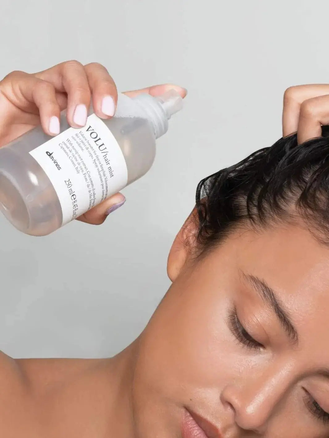 A person applies Davines VOLU Volumizing Hair Mist from a clear spray bottle directly onto their damp hair roots against a grey background | Twentyseven Toronto