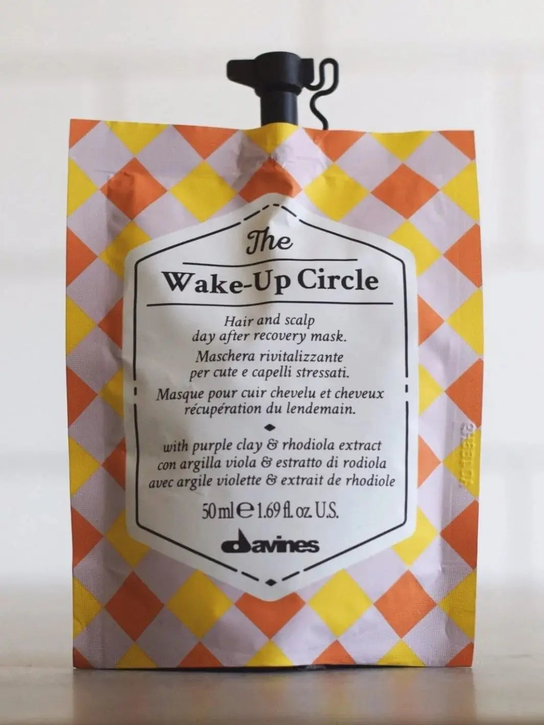 A colorful, diamond-checkered pouch of Davines The Wake-Up Circle of Chronicles Hair Mask stands on a flat surface against a white background | Twentyseven Toronto