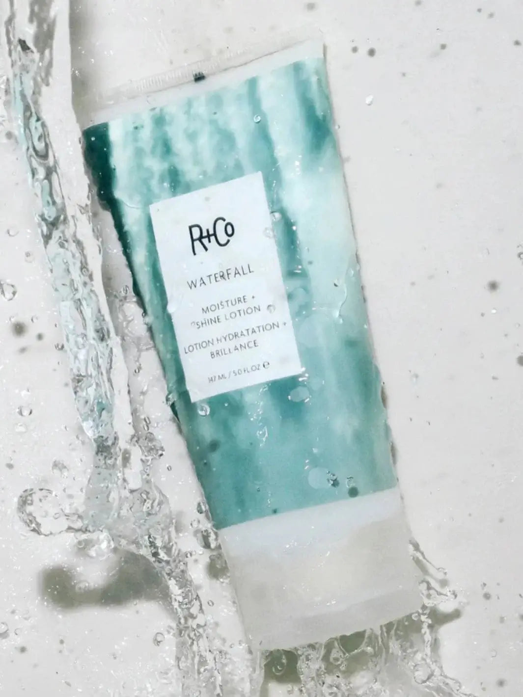A tube of R+Co WATERFALL Moisture + Shine Lotion lays flat against a white surface as fresh water splashes over it | Twentyseven Toronto